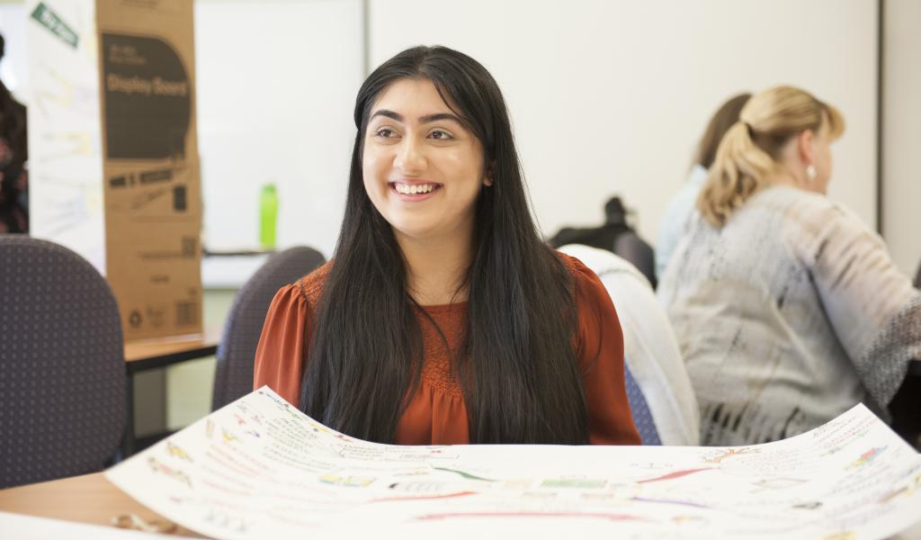 Seated, smiling female student sits in front a class project