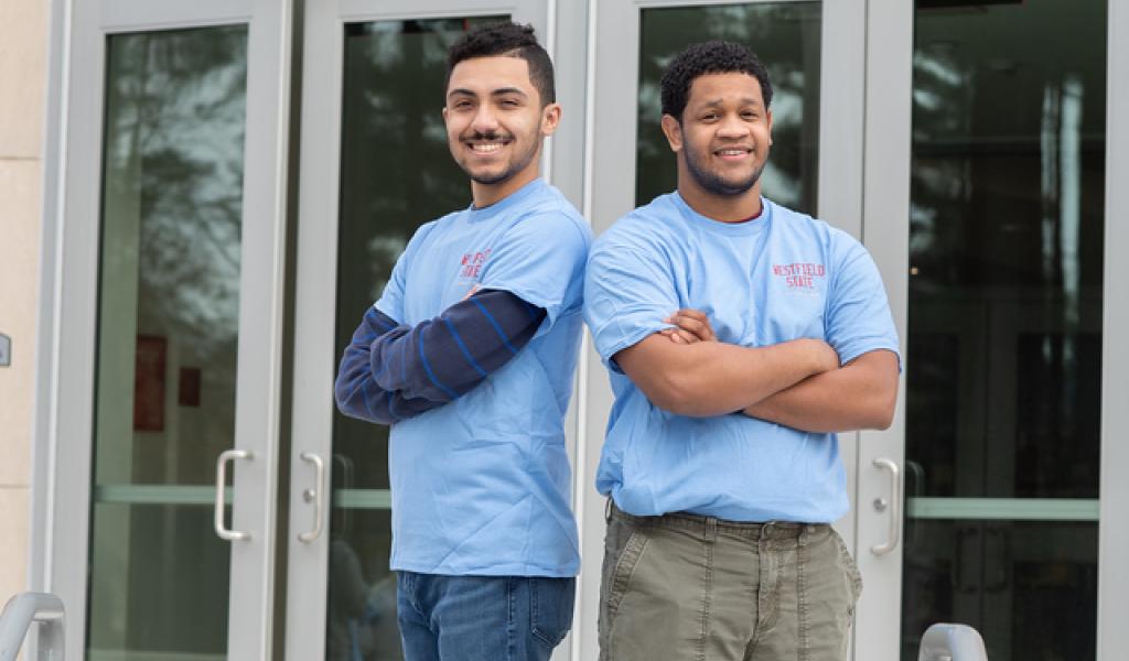 Two students in front of Parenzo wearing blue WSU shirts back to back with arms crossed.