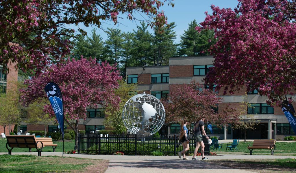 Two students walk in front of the campus globe surrounded by pink flowering trees on a bright spring day.