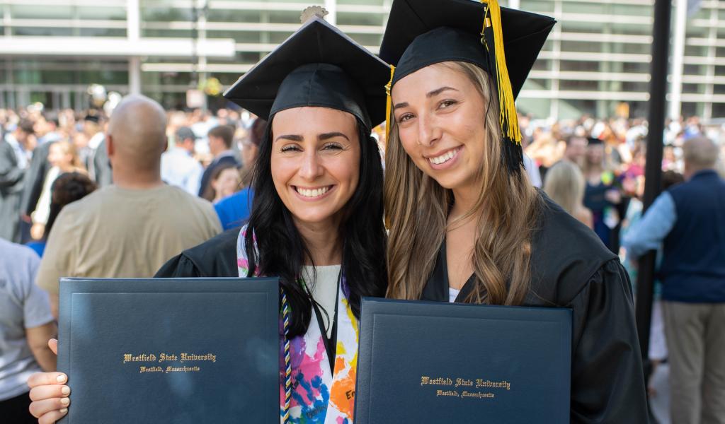Two graduates in caps and gowns smiling and holding diplomas at commencement.