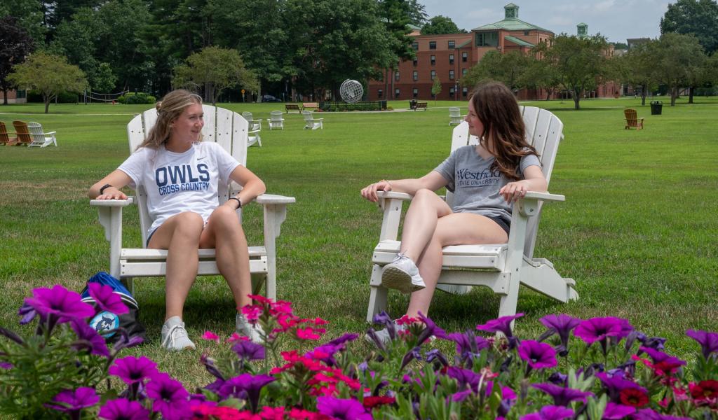 Two students sitting in Adirondack chairs surrounded by flowers on the campus green.