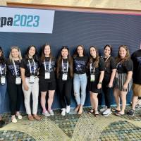 A photo of Andrew Allard, Nicole Clemente, Joelle Encarnacion, Melinda Hurteau, Giselle Lemus Tejada,  Kadeja Miller, Jennifer Petrucci, Taylor Saimeri, Mark Sigwart, and Hailey Whipple (not in order), smiling in front of a dark-blue backdrop AAPA 2023 poster.