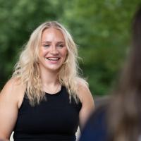 Jessica Gardner, a blond young woman in a black tanktop, smiles at a group of students blurred and in the corner of the left-hand side of the photo.