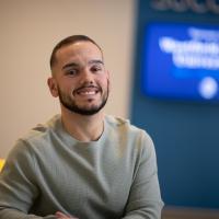 Nate Ferreira, sitting on a yellow chair with a blue and white background behind him that is out of focus.