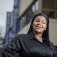 Ketia Valme, an English major and class of 2024, wears a black, long-sleeved shirt and smiles before the Ely center. 