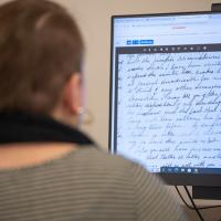 A close-up of a student participating in Douglass Day, 2024. Their back is turned to the camera and is blurry, though they are wearing a gray sweater and dark blue scarf. They sit at a desktop computer, which shows historical documents from the 19th century as part of Douglass Day.