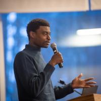 Zul Manzi, a Tanzanian-American content creator, is standing in front of a blue poster of trees. He is dressed in a long-sleeve, blue sweater and holds a microphone in his hand. He's speaking to a crowd of people who've gathered to watch his original show, "The Matimbulas".