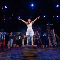 Westfield State's production of "Peter and the Starcatcher". A lone student wearing a blue dress is standing on top of a wooden chest on stage. Their hands and arms are splayed out above their head, at their sides. Several other actors surround them as they perform.