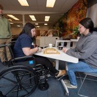 Occupational therapy students. Two sit across from each other on a white table and play jingo. Another stands by and watches.