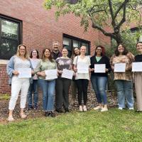 Newly graduated students from the Masters of Social Work program. They're standing with their certificates beside a tree and a brick building on campus.