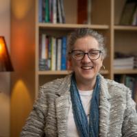 Dr. Kim Tobin, Professor of the Criminal Justice Department. She is sitting in her office and is seated before a bookcase. She wears a gray jacket and blue scarf.