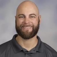 Alum Jake Suval. He is wearing a gray polo shirt and is posed in front of a professional, gray background.