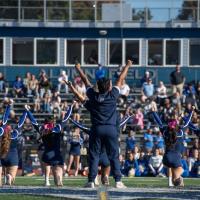 The University's cheerleading team performing in front of a crowd for this year's Homecoming weekend. Owls in blue uniforms face the crowd on bleachers and raise their hands in the air.